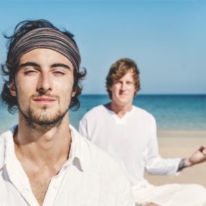 Two young men sitting cross-legged and meditating on the beach.