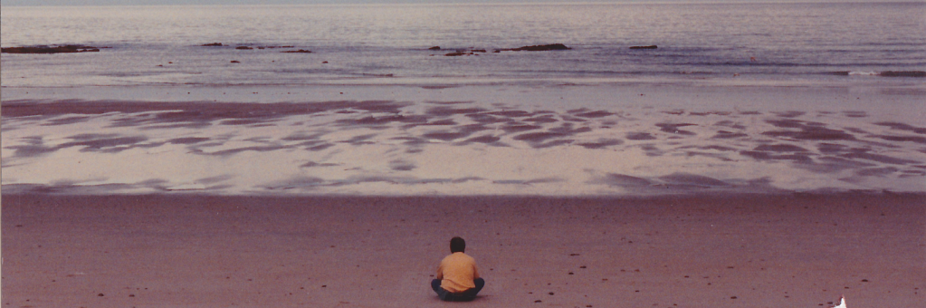 Image of CW sitting cross-legged, meditating on a beach, while watching the sun set.
