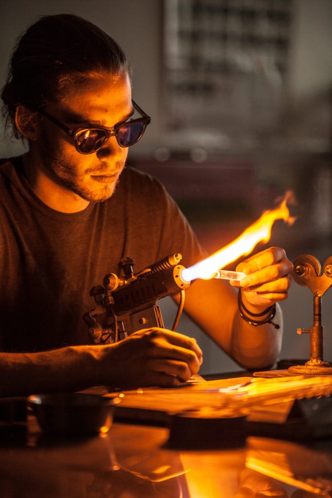 Glassblowing artisan working on glass tubes with a blow-torch flame.