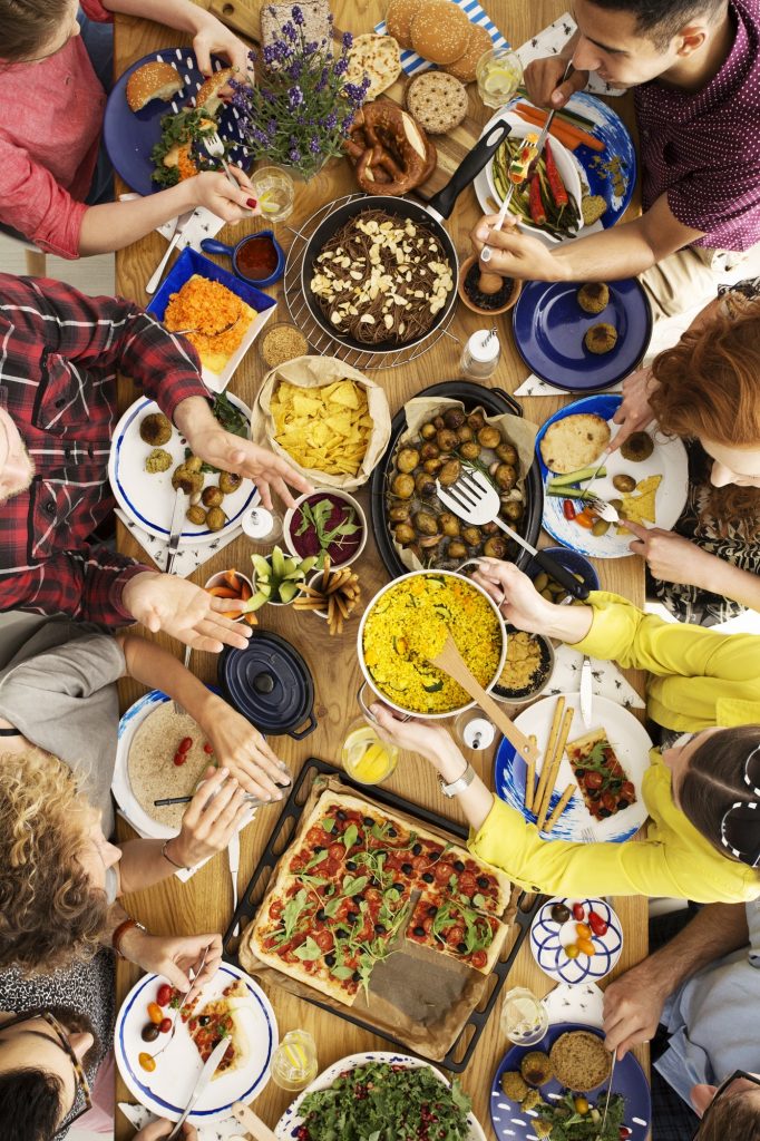 Overhead view of a group of friends sharing a meal. Table is laden with food. People are passing heaping-full platters and bowls around. Diverse group of ethnicities and gender identities.