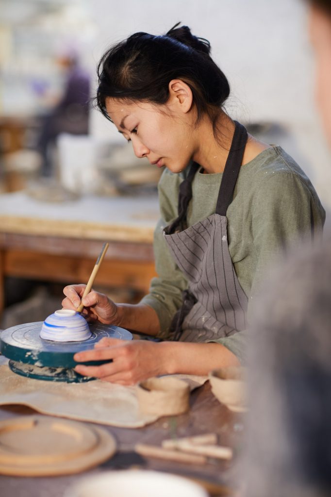 Young woman in apron, sitting at workshop table painting a clay bowl on a turntable.